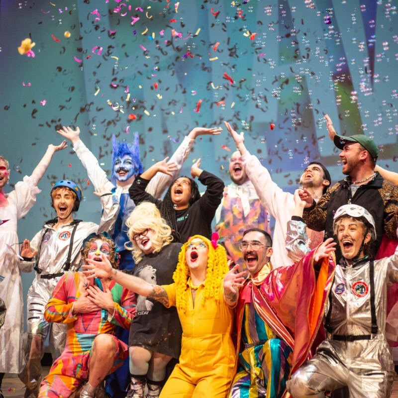 A group of people wearing colourful outfits stand in a bunch with their arms in the air, celebrating as confetti falls on them