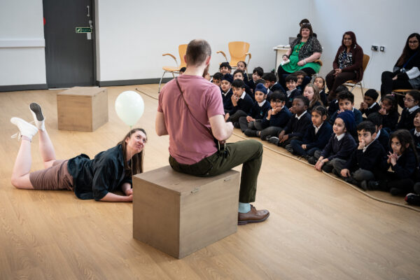 In front of an audience of children, 1 person sits on a box and from the back we can see they are playing a ukulele, another performers lies next to them, looing up, watching them play
