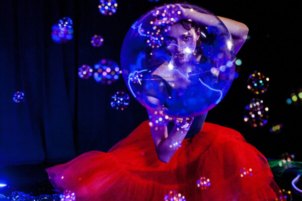 A dancer crouches, holding a giant bubble between her hands, and looks through the bubble directly at the camera lense