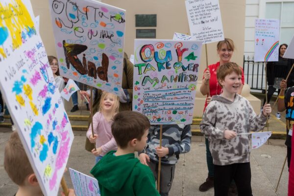 A group of children have banners and are marching outside a large white building.